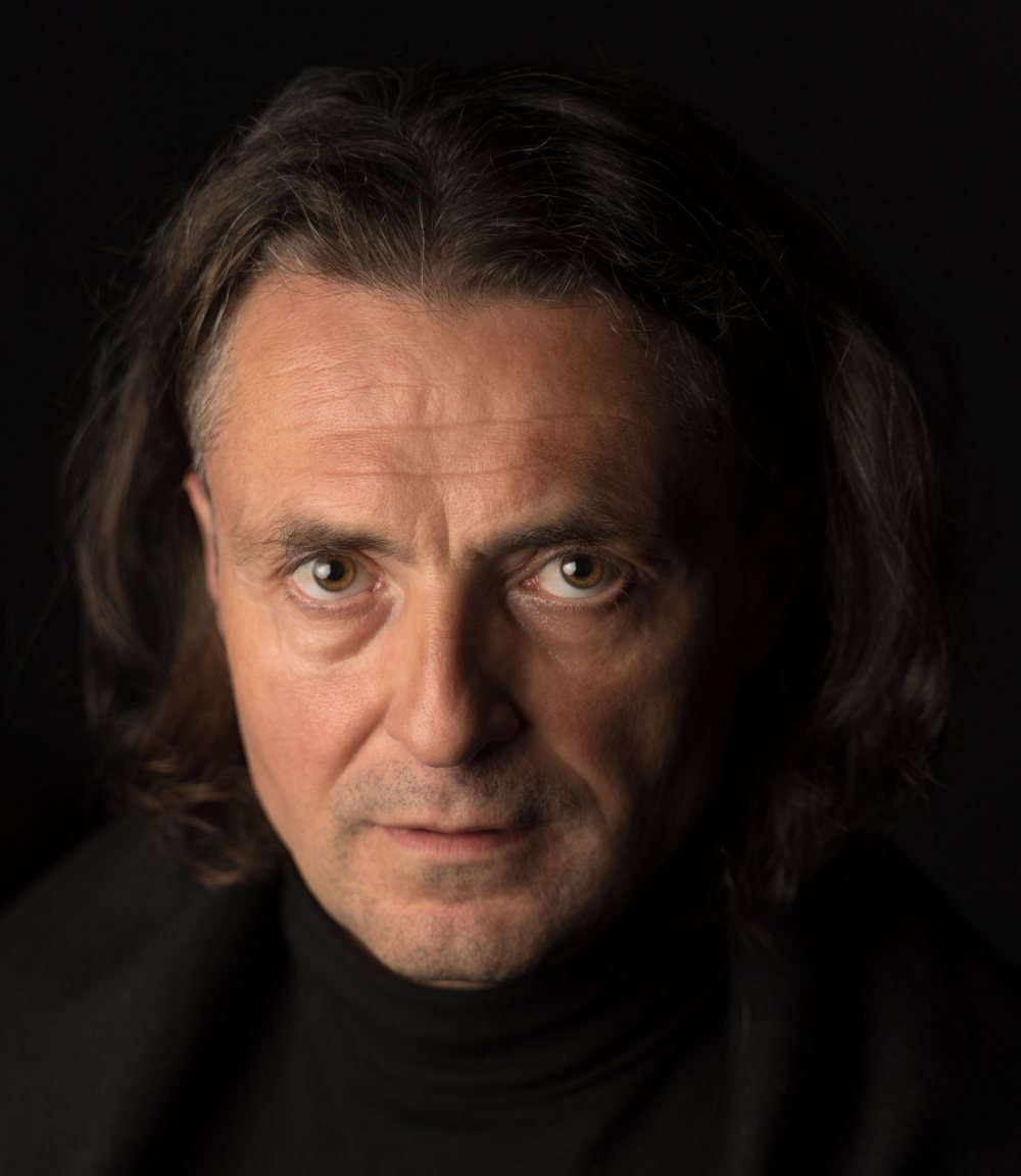 Boris Baňas, portrait on black: long hair, black turtleneck, looking straight at the camera.