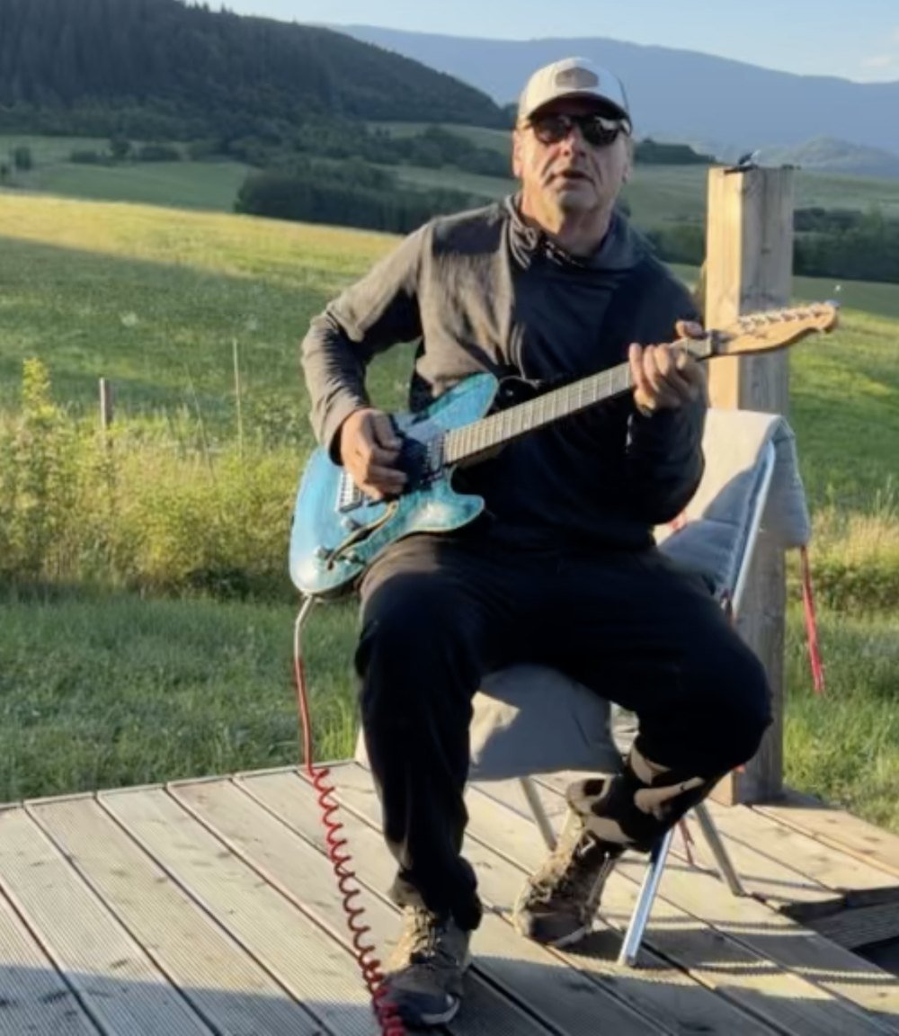 Peter Karetka on a wooden deck, cap and sunglasses, green hills behind.